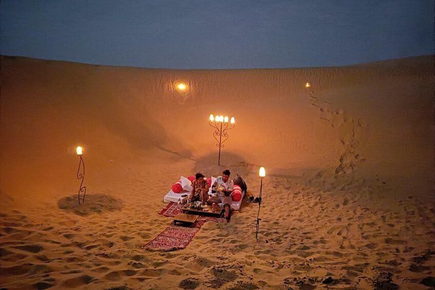 A desert scene with a group of people dining on a table set up on the sand, surrounded by desert dunes and illuminated by candlelight. 3