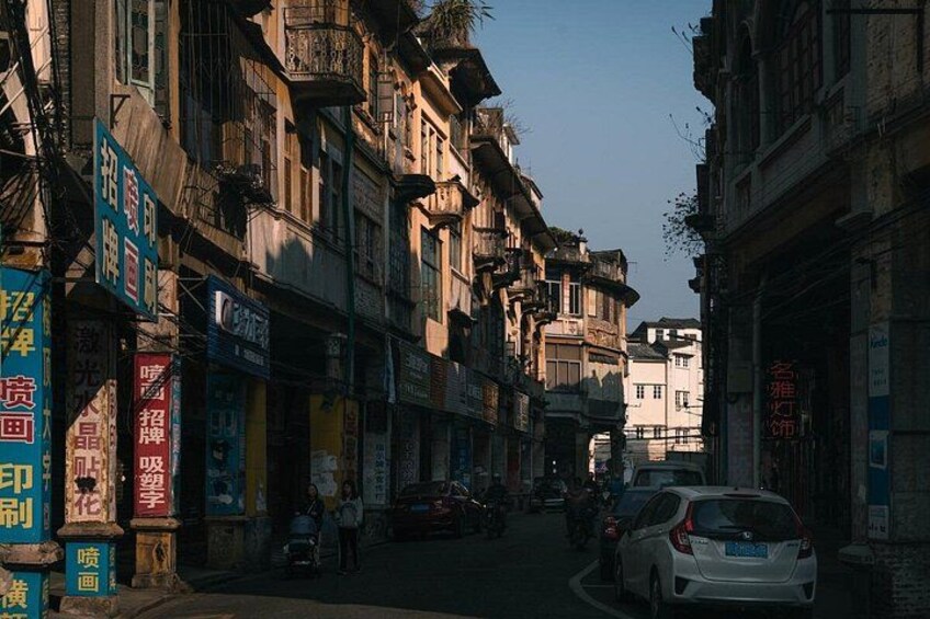 A narrow street lined with multi-story buildings, some with balconies, and various signs and advertisements. 1