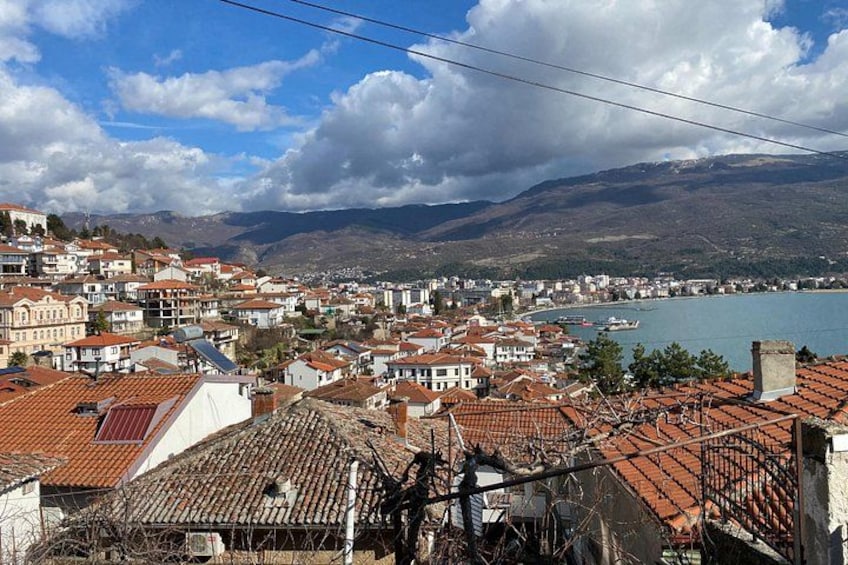 A coastal town with terracotta-roofed buildings, a body of water, and mountains in the background. 3