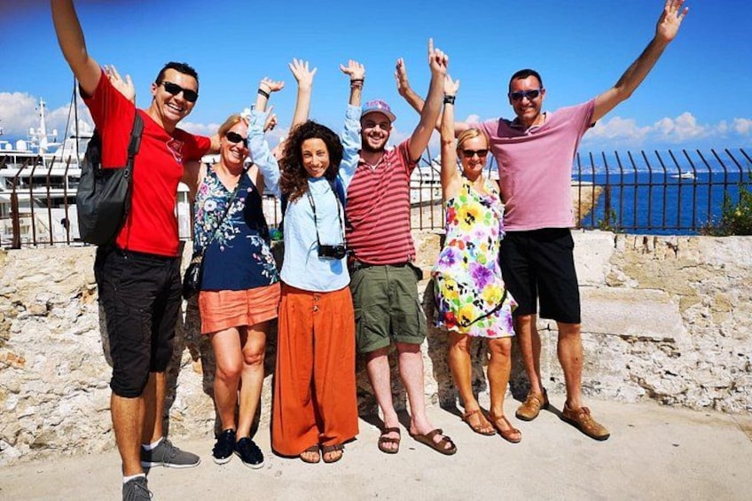A group of people standing on a rocky shore with their arms raised, a clear sky, and a body of water in the background. 3