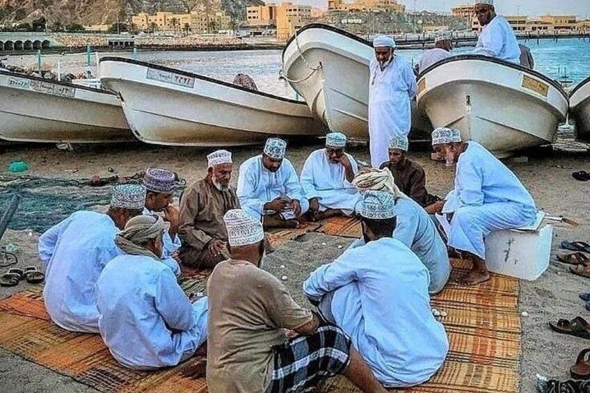 Un grupo de hombres sentados en esteras junto a la orilla, con barcos al fondo. 3