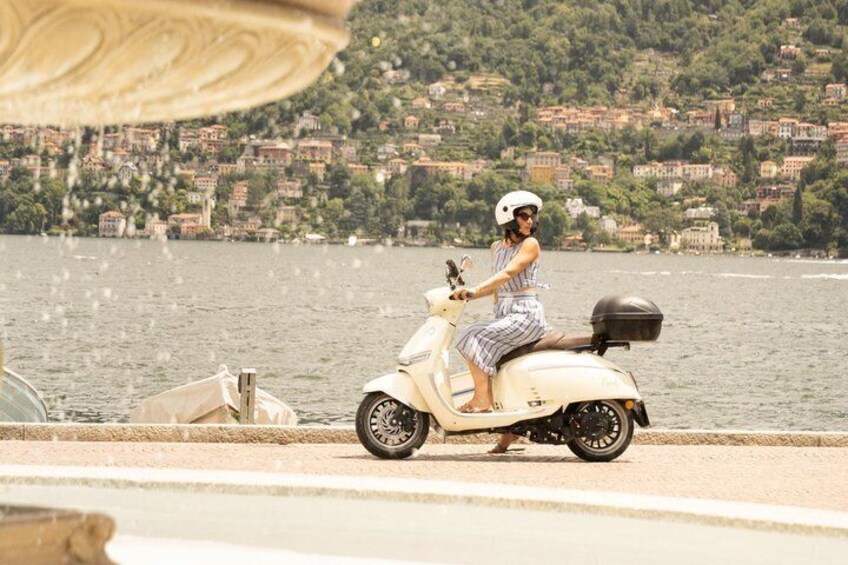 A person riding a white scooter by a waterfront with buildings in the background. 2