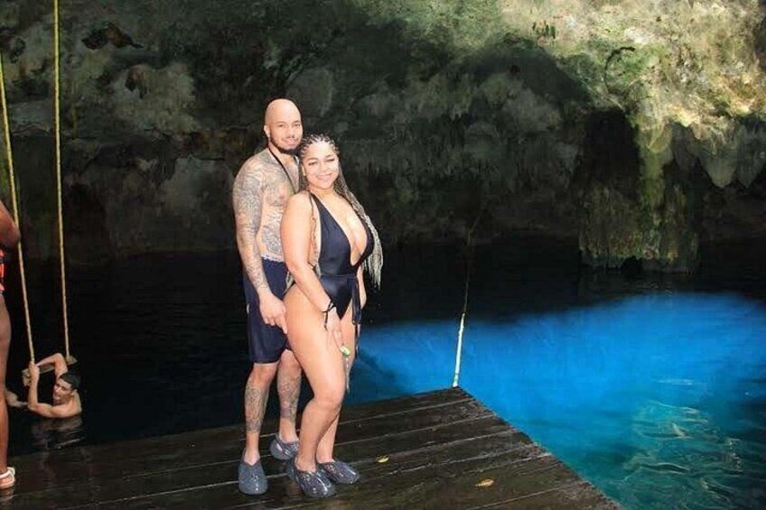Ein Paar steht auf einem hölzernen Steg über einem blauen Becken, im Hintergrund eine höhlenartige Landschaft. 2