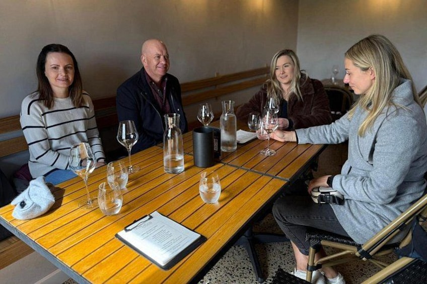 A group of people seated around a wooden table with wine glasses and bottles. 1