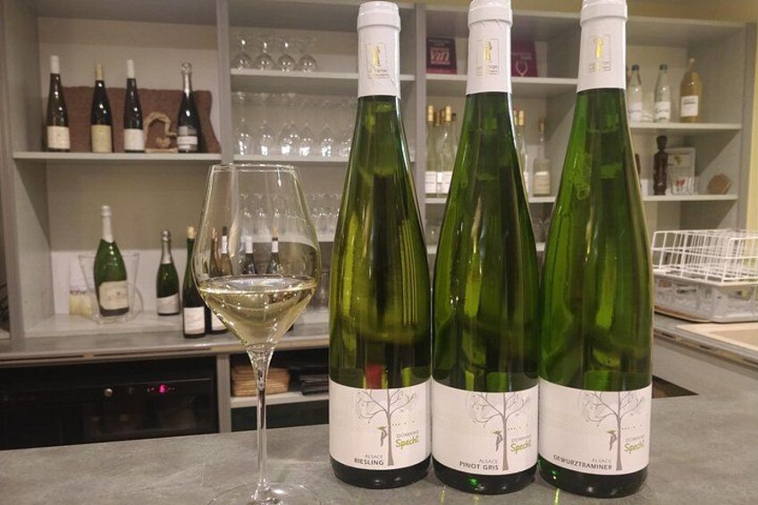 Tre flaskor vin med vita etiketter och ett vinglas fyllt med vitt vin står på en bänk. 2