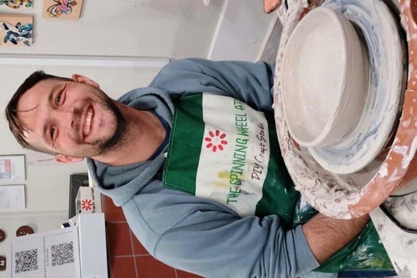 A person wearing a green apron with 'The Spinning Wheel' text, working with pottery on a wheel. 2