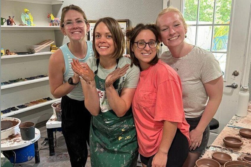 Four people in a pottery workshop with shelves of pottery and clay materials. 1