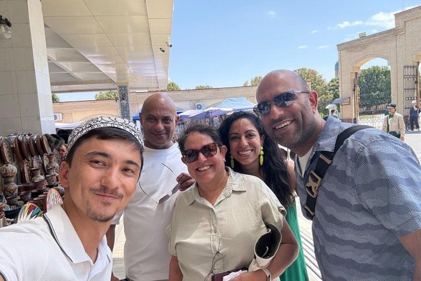 A group of people posing for a photo in an outdoor setting with a clear sky, a building with an archway, and a market stall with various items. 3