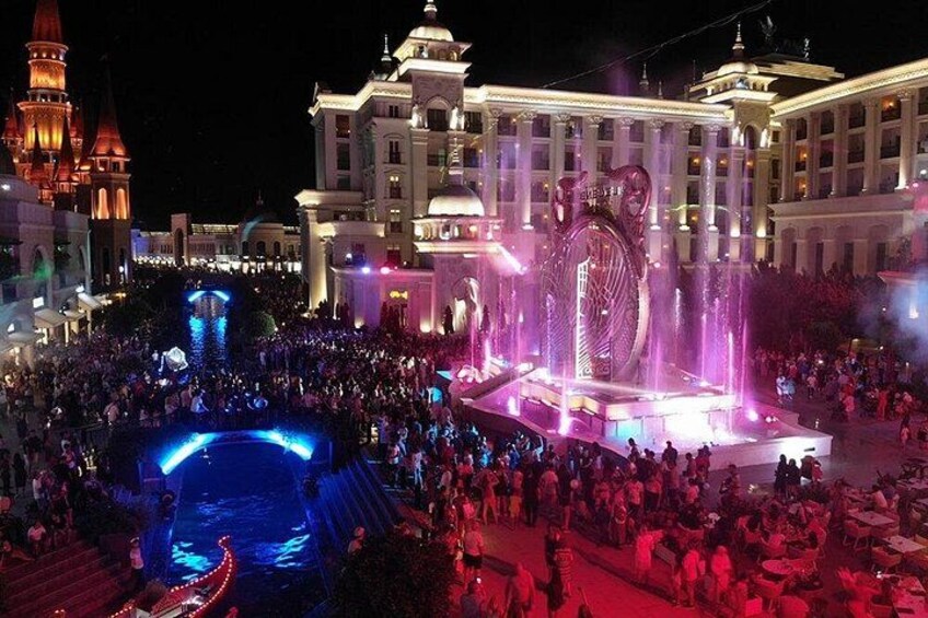 A large crowd gathered around a fountain with water jets and a large illuminated sculpture in the center. 3