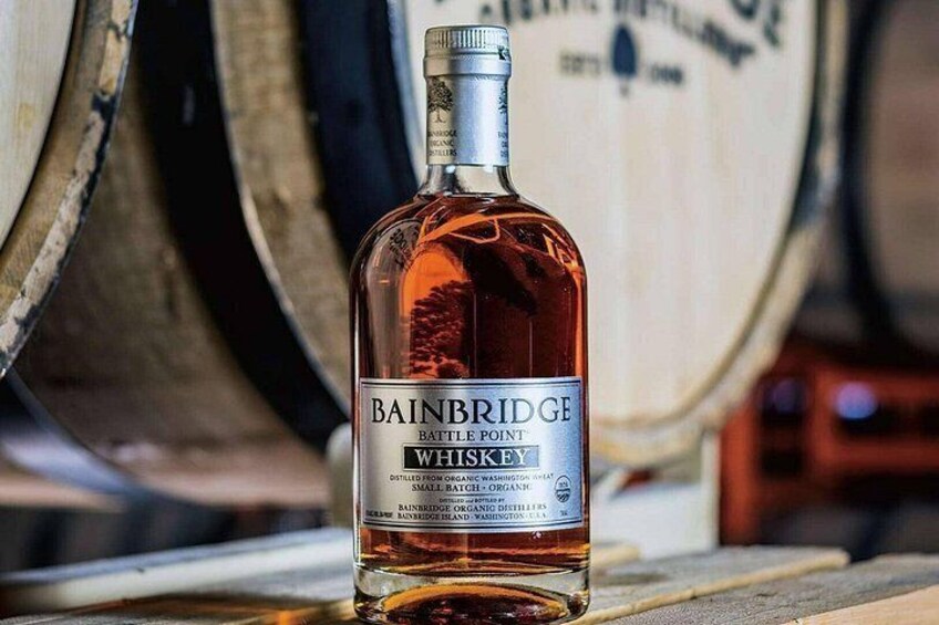 A bottle of Bainbridge Battelle Point Whiskey on a wooden surface with barrels in the background. 1