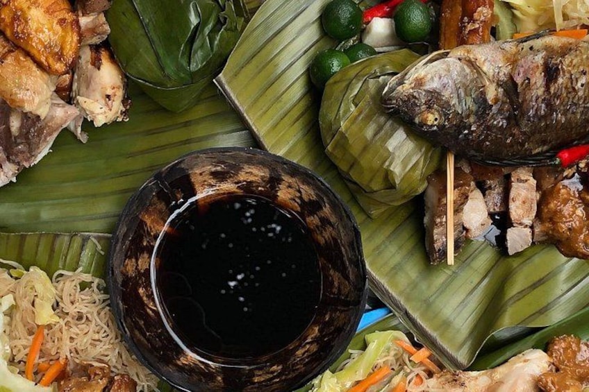 A variety of grilled fish and meats on banana leaves, a bowl of dark sauce, and some vegetables. 4
