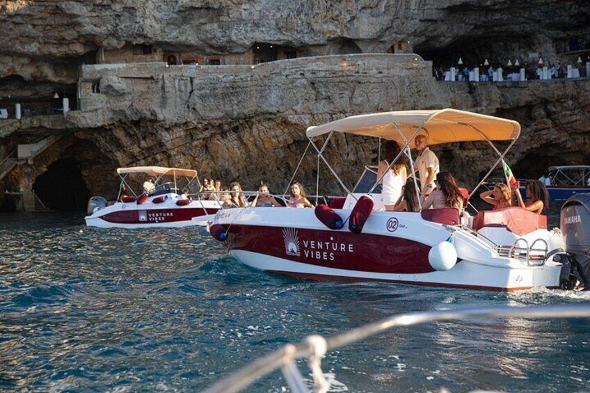 Two boats with 'Venture Vibes' branding, people on board, and a rocky cliff with a cave entrance in the background. 5
