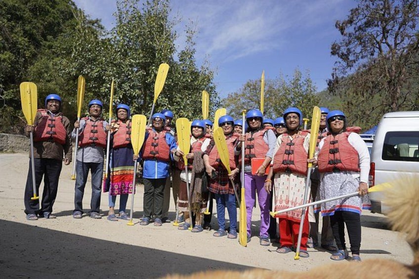 A group of people in life jackets and helmets, holding paddles, standing on a road with trees in the background. 3