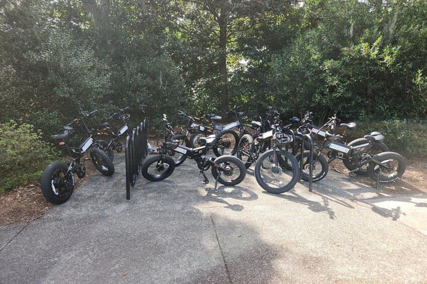 A row of parked bicycles and motorcycles in a shaded area with trees in the background. 2