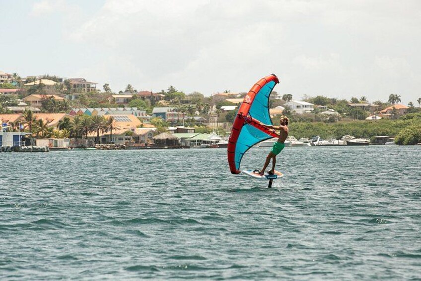 A person windsurfing on a body of water with a colorful sail, surrounded by a coastal town with buildings and palm trees. 4