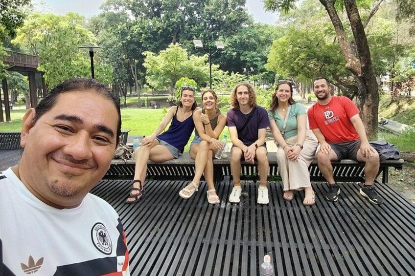 A group of people sitting on a bench in a park with trees and a wooden structure in the background. 4