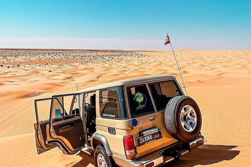 Un vehículo todoterreno color beige con una llanta de repuesto montada en la parte trasera, estacionado en un desierto arenoso con dunas al fondo. 5