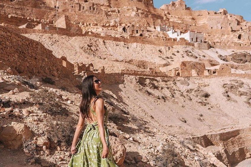 Una persona con un vestido verde de pie en un paisaje rocoso con ruinas antiguas al fondo. 1