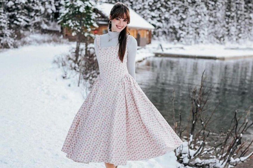 A person in a patterned dress standing by a snow-covered pond with trees and a wooden structure in the background. 2