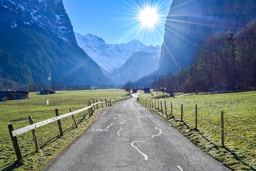 A paved road flanked by wooden fences, leading towards a mountainous landscape with a clear sky. 3