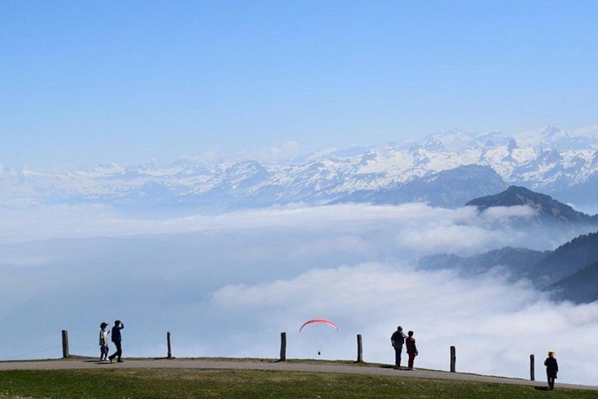 A group of people on a grassy hill overlooking a mountain range with snow-capped peaks and a paraglider in the distance. 2
