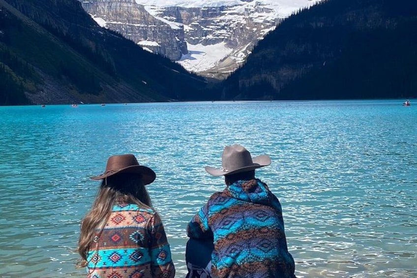 Twee personen met opvallende poncho's en hoeden, staand bij een turquoise meer met bergen op de achtergrond. 5