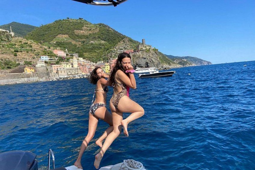 Dos personas en traje de baño en un barco cerca de una ciudad costera con edificios y una colina al fondo. 3
