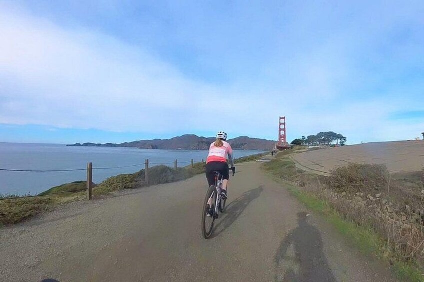 A cyclist on a paved path with a coastal view, a red bridge, and hills in the distance. 1
