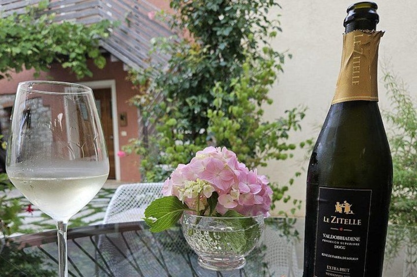 Eine Flasche Le Zitelle Wein, ein Glas Weißwein und eine rosa Hortensienblüte auf einem Tisch. 3