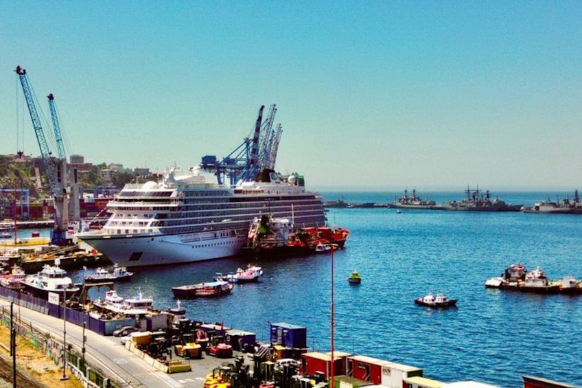 A large cruise ship docked at a port with cranes and smaller boats in the background. 1