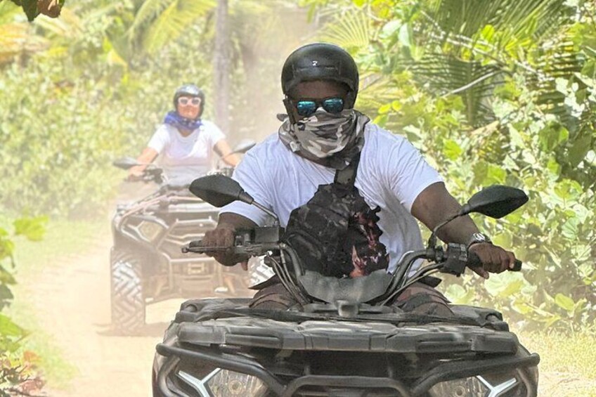 Two people on ATVs on a dirt path surrounded by greenery. 2
