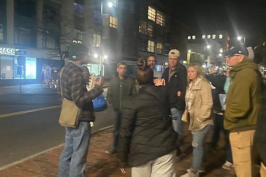 A group of people standing on a sidewalk at night, with buildings and a street in the background. 4
