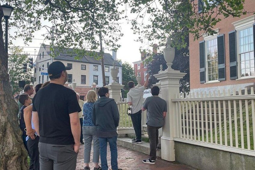 A group of people standing on a brick pathway, a white fence, and a building with a brick facade. 2