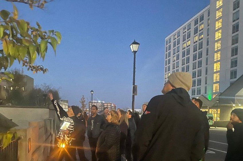 A group of people gathered on a city street at dusk, with buildings and a street lamp in the background. 1