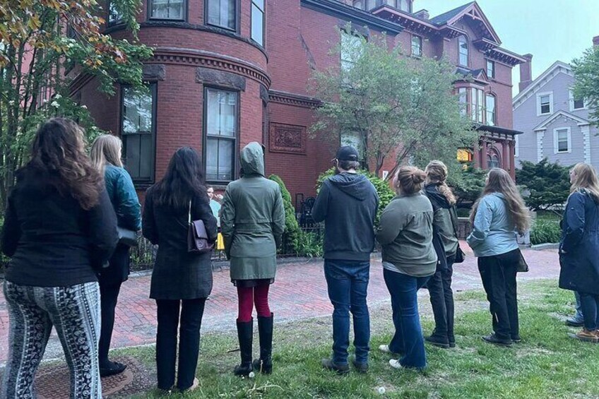 Un grupo de personas de pie sobre un camino de ladrillo frente a un edificio de ladrillo rojo con grandes ventanales. 4