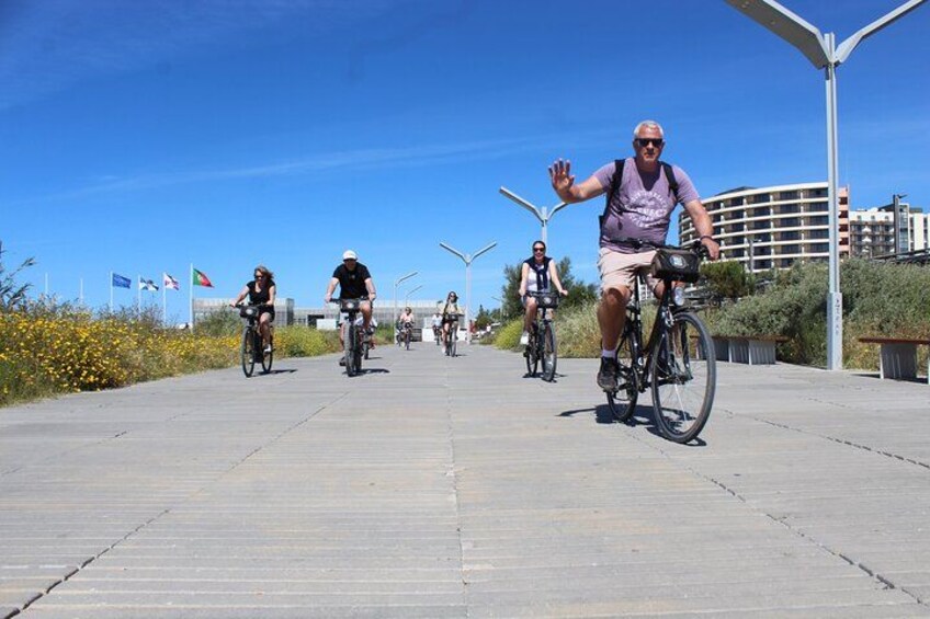 En grupp cyklister på en asfalterad stig, med flaggor och byggnader i bakgrunden. 2