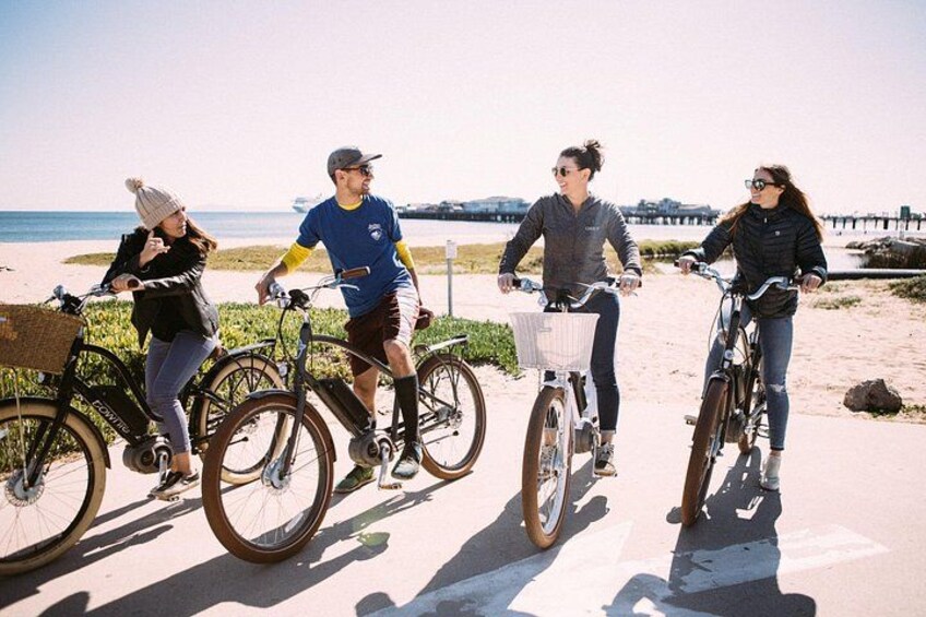 Four people riding bicycles on a paved path near a beach with a pier in the background. 2