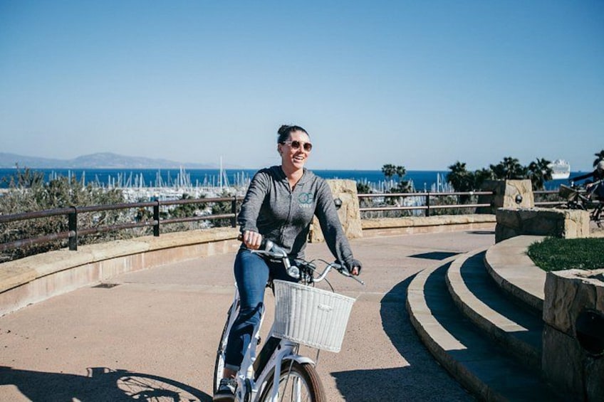 A person riding a bicycle with a basket on a paved path by the sea. 3