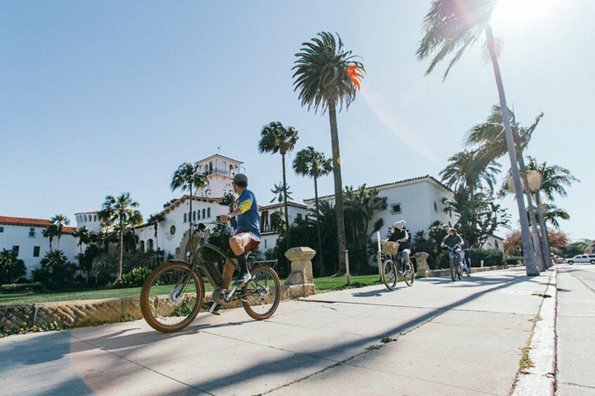 A sunny day with cyclists on a paved path, palm trees, and a white building with a red roof. 5
