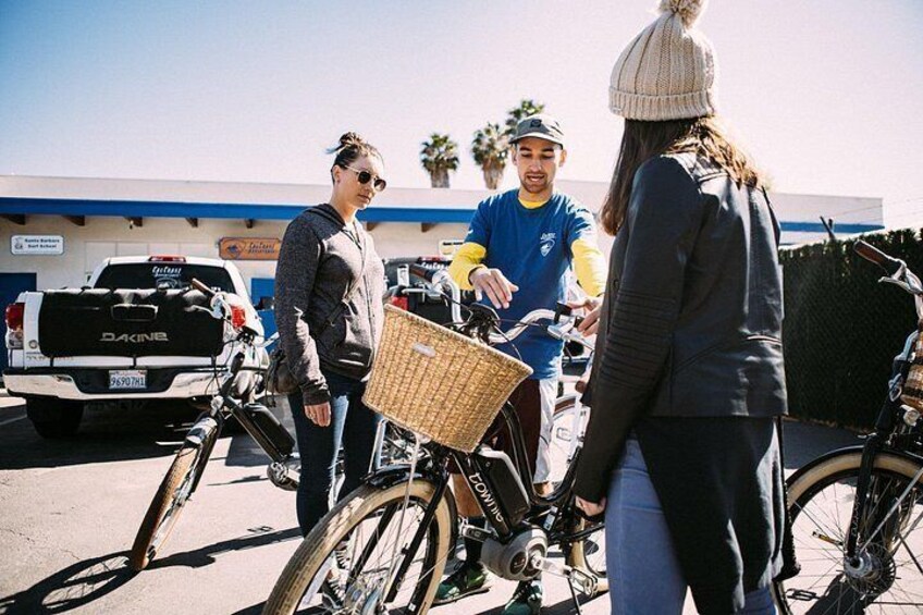 People standing around bicycles in a parking lot with a truck and a building in the background. 2
