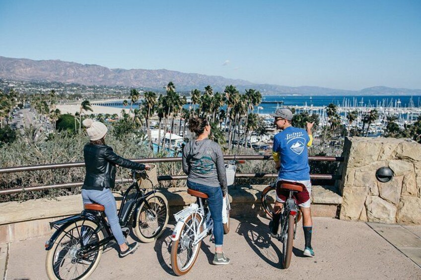 Three people on bicycles, a coastal view with palm trees, and a marina in the background. 1