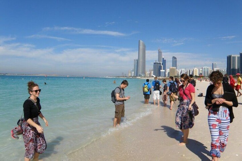 En strand med folk som promenerar, en stadsbild i bakgrunden och klart, blått vatten. 3
