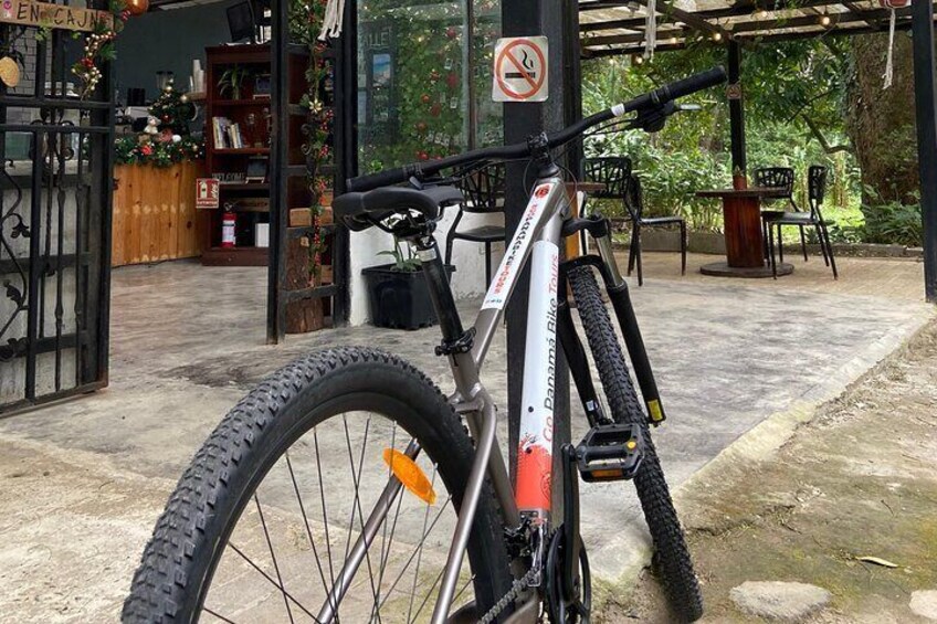 A mountain bike parked in a covered outdoor area with a wooden table and chairs, surrounded by greenery. 3
