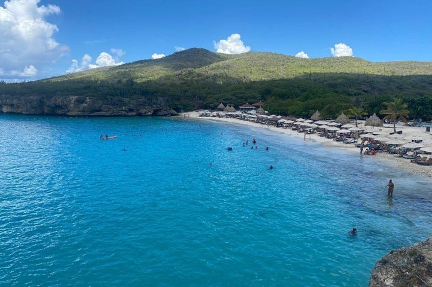 A beach with clear turquoise water, people swimming, and a hilly landscape in the background. 2