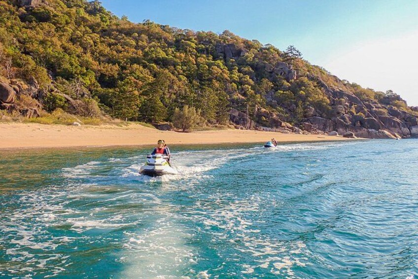 A person jet-skiing on a clear blue sea with a forested hill in the background. 1
