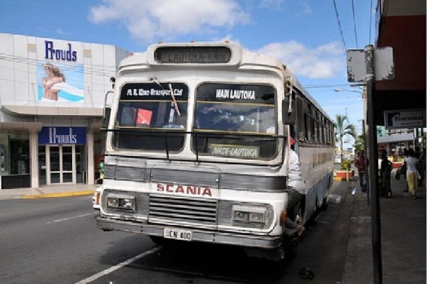 A white Scania bus with 'MARI LAUTOKA' on the front, parked on a street with a 'Rouds' store in the background. 2
