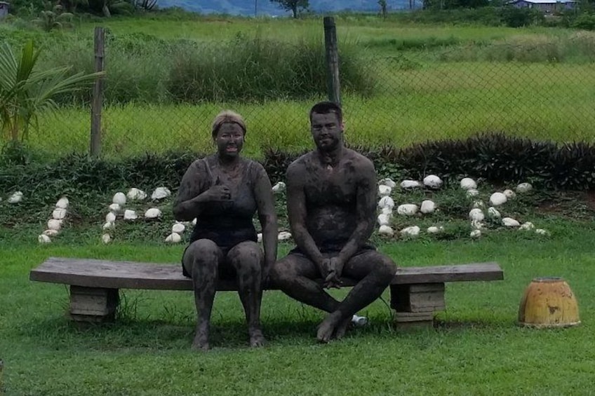 Two people sitting on a bench in a grassy area with a fence and scattered objects in the background. 5