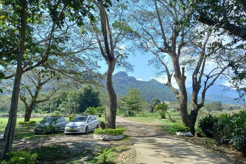 A paved path, two parked cars, and trees in a rural setting with mountains in the background. 4