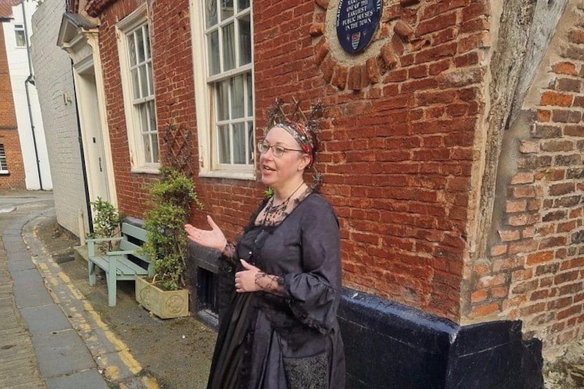 A person in historical costume standing in front of a brick building with a plaque. 4