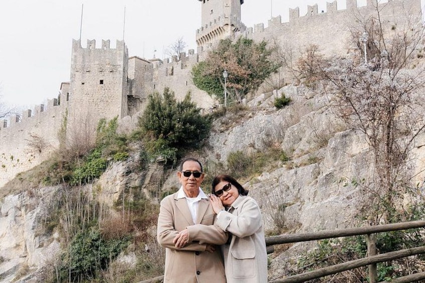 A man and a woman standing in front of a historic castle with a stone wall and a wooden fence. 1